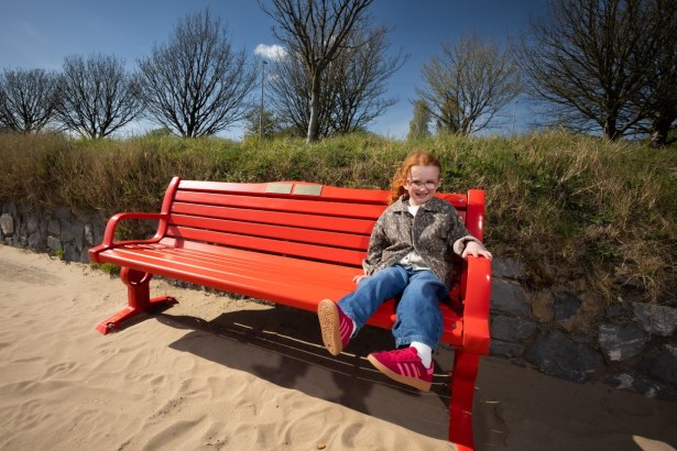 Esme Mariani, with red hair and glasses, smiling as she sits on a bright red British Heart Foundation bench on the sand at Swansea Beach