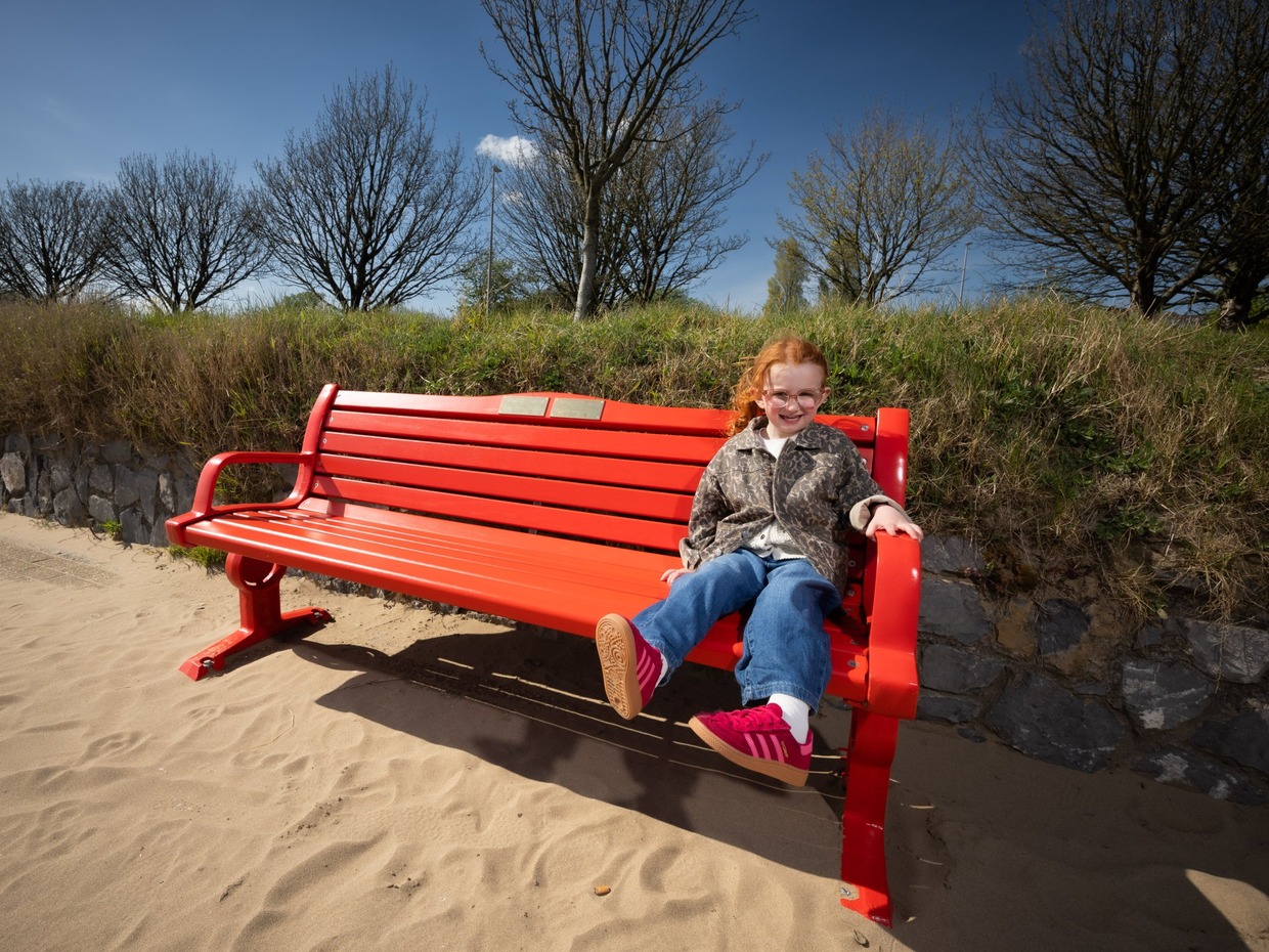 Esme Mariani, with red hair and glasses, smiling as she sits on a bright red British Heart Foundation bench on the sand at Swansea Beach