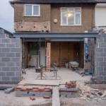 The rear of a semi-detached house mid-extension, with new blockwork walls, a steel beam, acrow props, a wheelbarrow and stacked bricks on the ground
