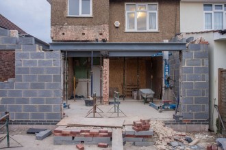 The rear of a semi-detached house mid-extension, with new blockwork walls, a steel beam, acrow props, a wheelbarrow and stacked bricks on the ground