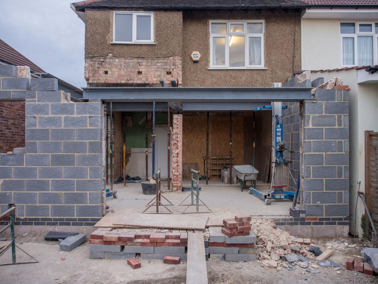 The rear of a semi-detached house mid-extension, with new blockwork walls, a steel beam, acrow props, a wheelbarrow and stacked bricks on the ground
