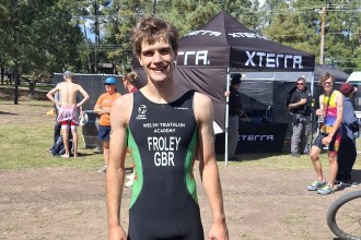 Iwan Froley smiling in a black and green Welsh Triathlon Academy race suit marked FROLEY GBR, with an XTERRA tent and pine trees behind him