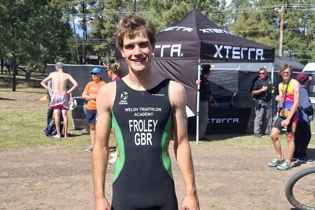 Iwan Froley smiling in a black and green Welsh Triathlon Academy race suit marked FROLEY GBR, with an XTERRA tent and pine trees behind him