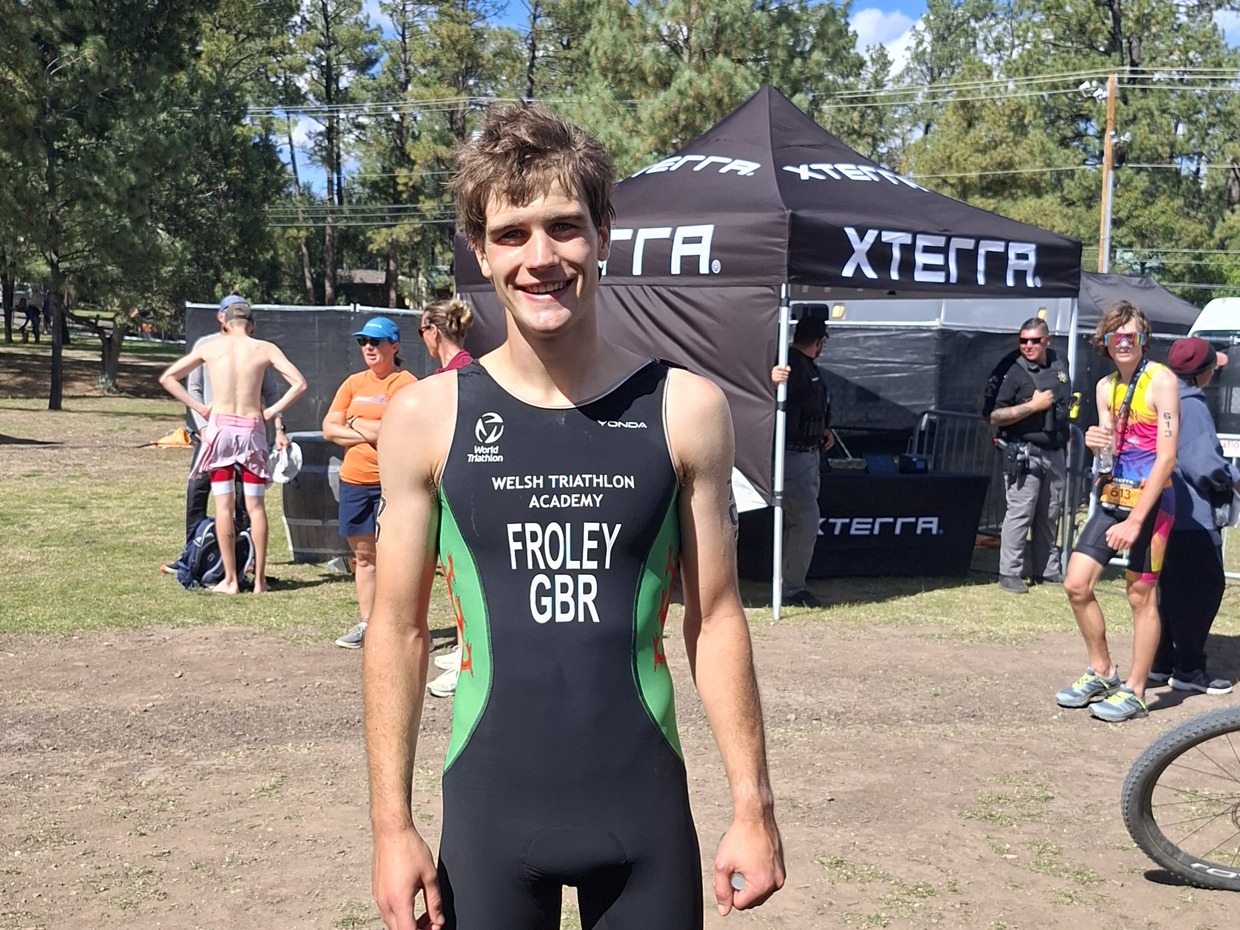 Iwan Froley smiling in a black and green Welsh Triathlon Academy race suit marked FROLEY GBR, with an XTERRA tent and pine trees behind him