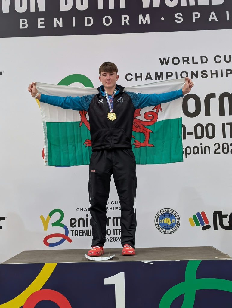 Jacob Wyatt stands on the top step of the podium in Benidorm holding a Welsh flag behind him, with a gold medal round his neck