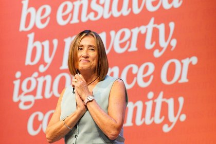 Jane Dodds with her hands clasped on stage in front of a red backdrop reading be enslaved by poverty, ignorance or conformity