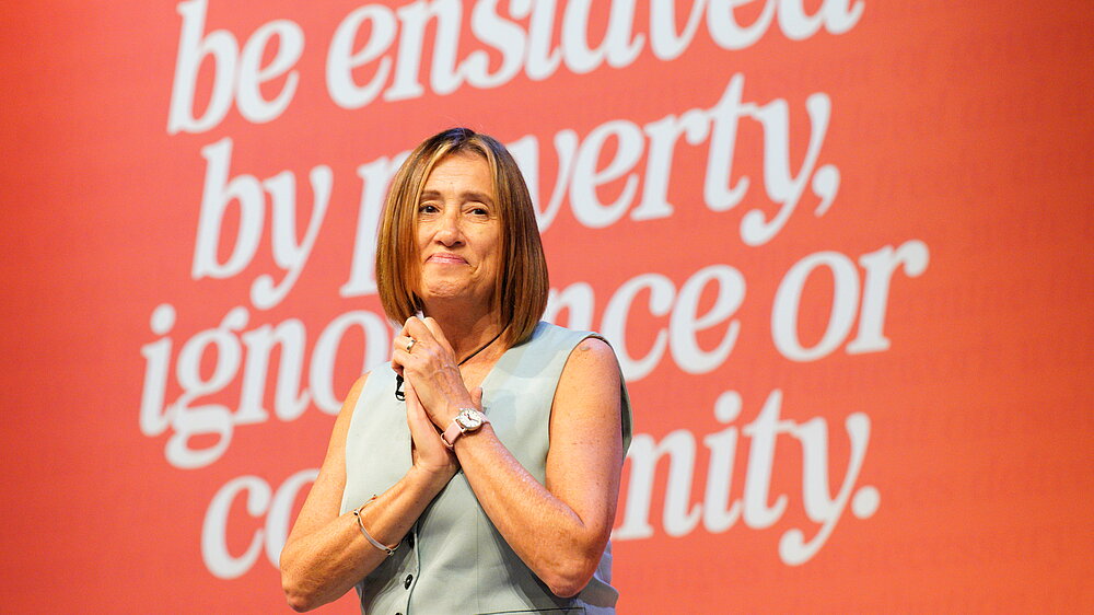 Jane Dodds with her hands clasped on stage in front of a red backdrop reading be enslaved by poverty, ignorance or conformity