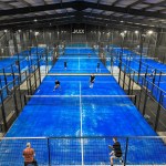 Nine bright blue indoor padel courts under a steel roof with the Jaxx sign on the far wall and players mid-game