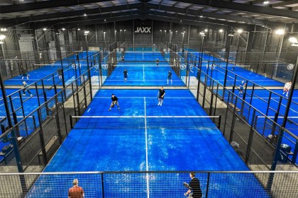 Nine bright blue indoor padel courts under a steel roof with the Jaxx sign on the far wall and players mid-game
