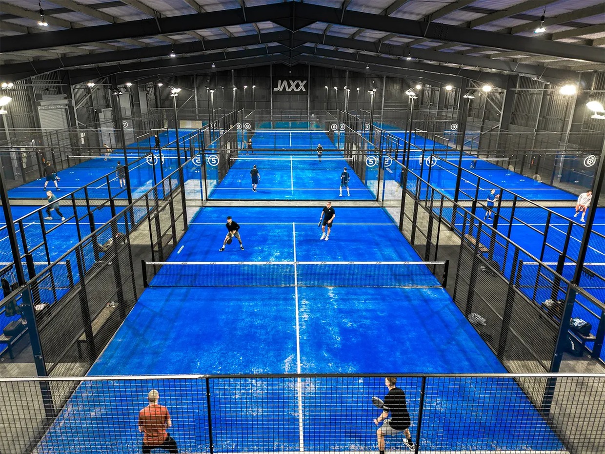 Nine bright blue indoor padel courts under a steel roof with the Jaxx sign on the far wall and players mid-game