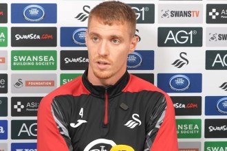 Swansea City midfielder Jay Fulton, in a red and black training top, speaking at a press conference in front of a sponsor board