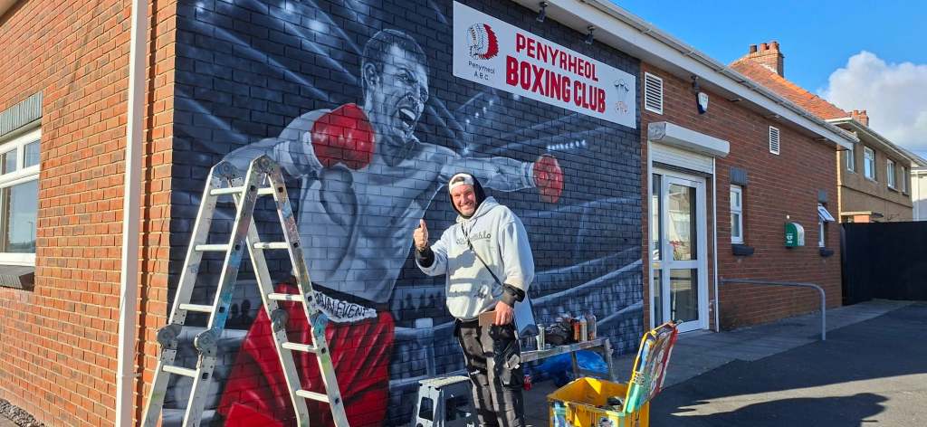 Artist Jenks gives a thumbs-up beside his mural of Colin Jones on the wall of Penyrheol Boxing Club, with a stepladder and spray cans nearby.