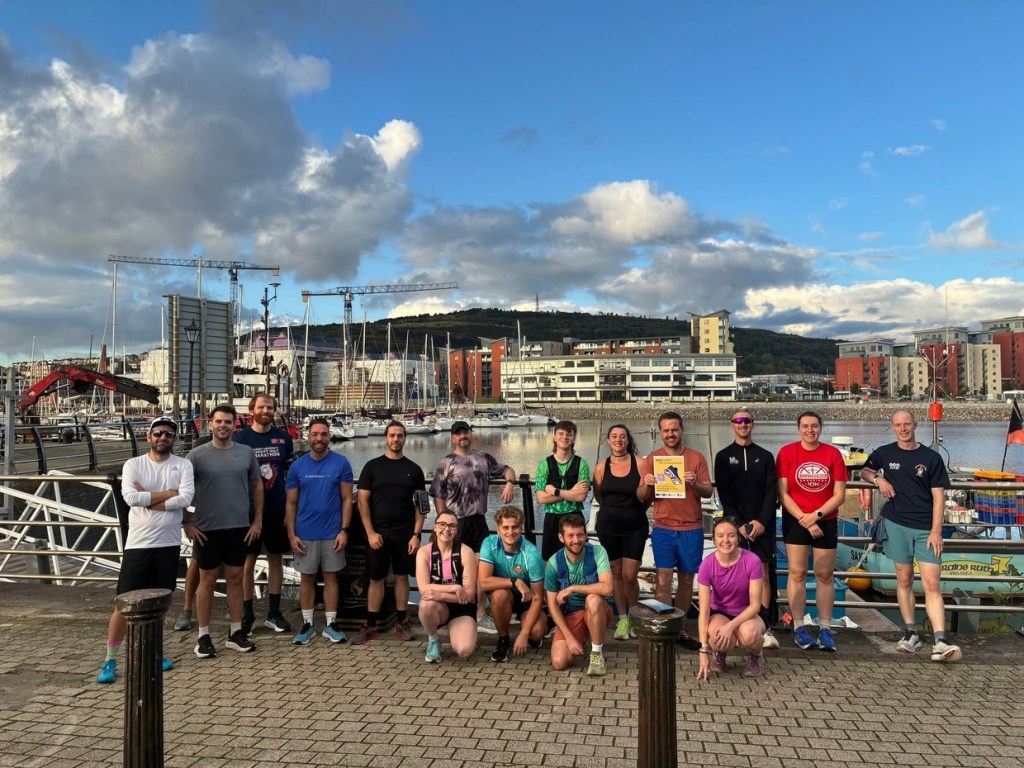 A running club group posing beside Swansea Marina with Kilvey Hill behind, one runner holding a JogOn poster