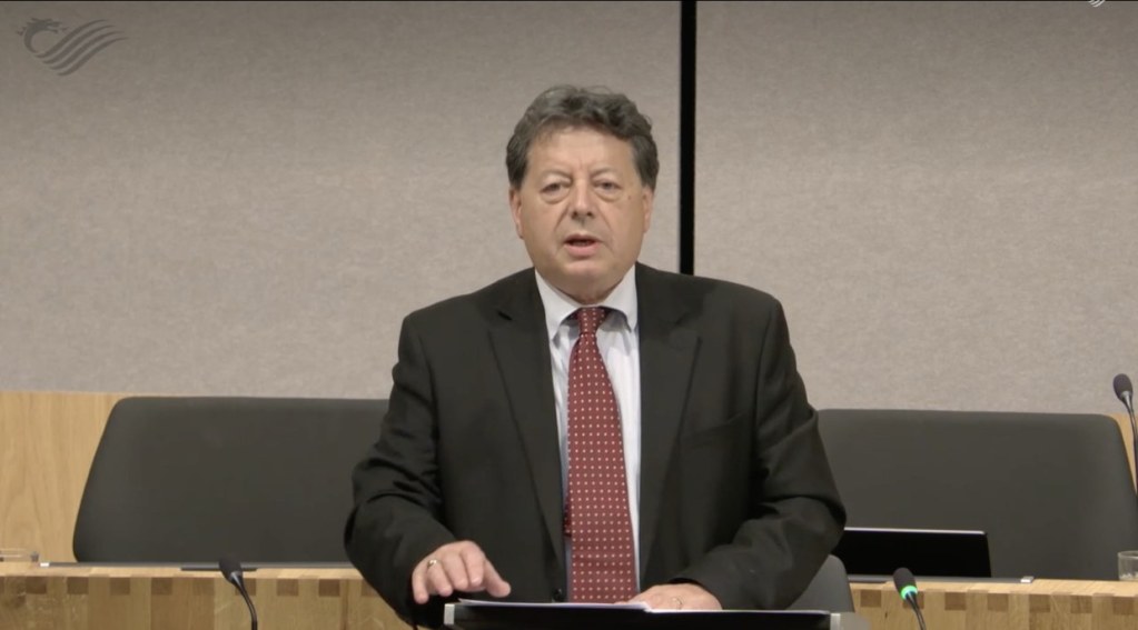 John Davies, in a dark suit and red spotted tie, speaking at a lectern in the Senedd chamber