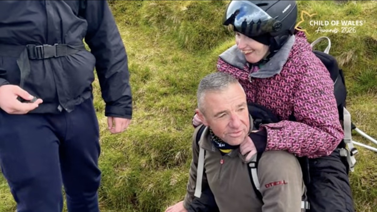 John Jenkins kneeling on the grassy hillside with his daughter Leah, in a helmet, laughing on his back in an adapted carrier