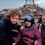 Macsen, Leah in a helmet and pink jacket, and John Jenkins smiling beside the summit marker on Pen y Fan
