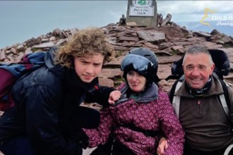 Macsen, Leah in a helmet and pink jacket, and John Jenkins smiling beside the summit marker on Pen y Fan