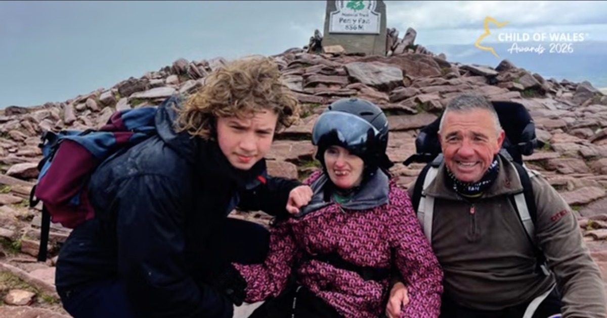 Macsen, Leah in a helmet and pink jacket, and John Jenkins smiling beside the summit marker on Pen y Fan