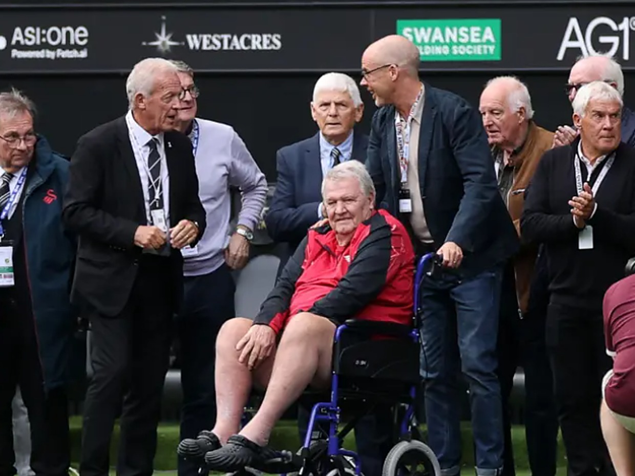 John Toshack, seated in a red and black jacket, surrounded by members of Swansea City's 1981 team pitchside at the Swansea.com Stadium