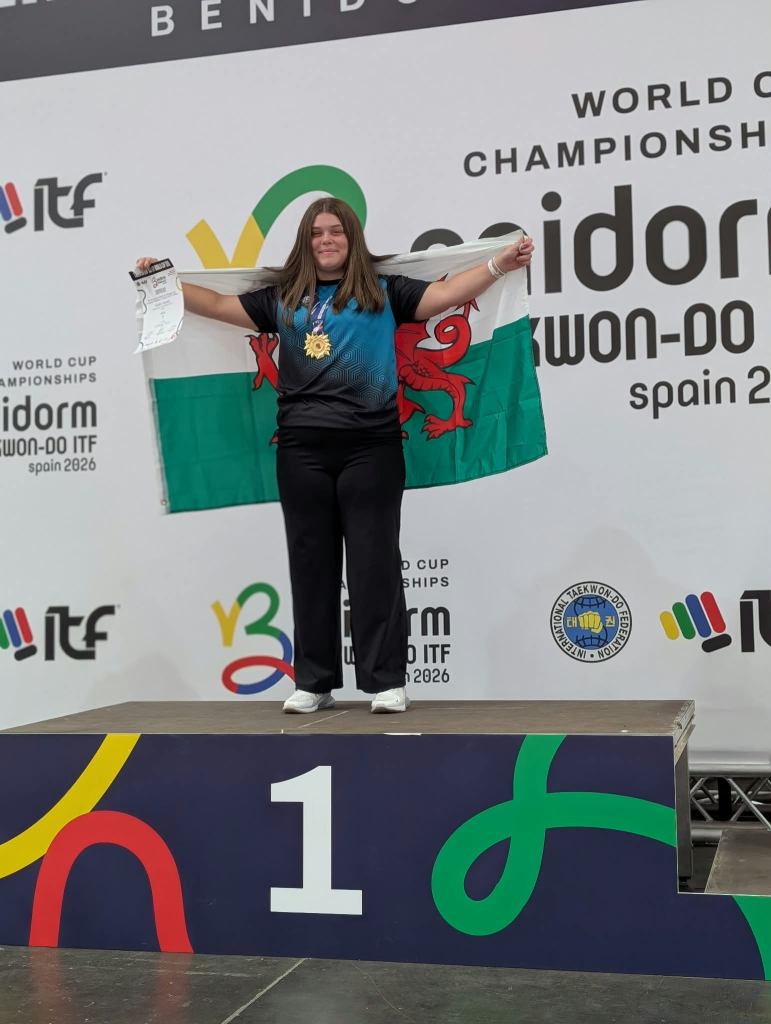 Kelsey Davies stands on the top step of the podium in Benidorm with a Welsh flag, holding her certificate and wearing a gold medal