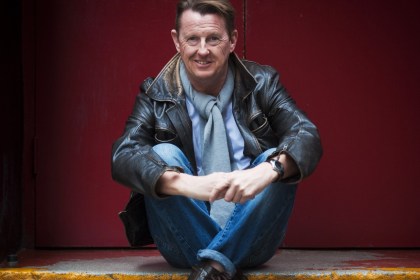 Kevin Kennedy in a brown leather jacket, grey scarf and jeans, sitting on a step in front of a red door and smiling