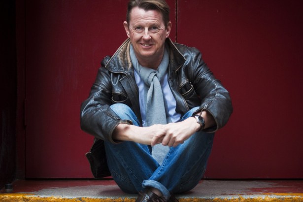 Kevin Kennedy in a brown leather jacket, grey scarf and jeans, sitting on a step in front of a red door and smiling