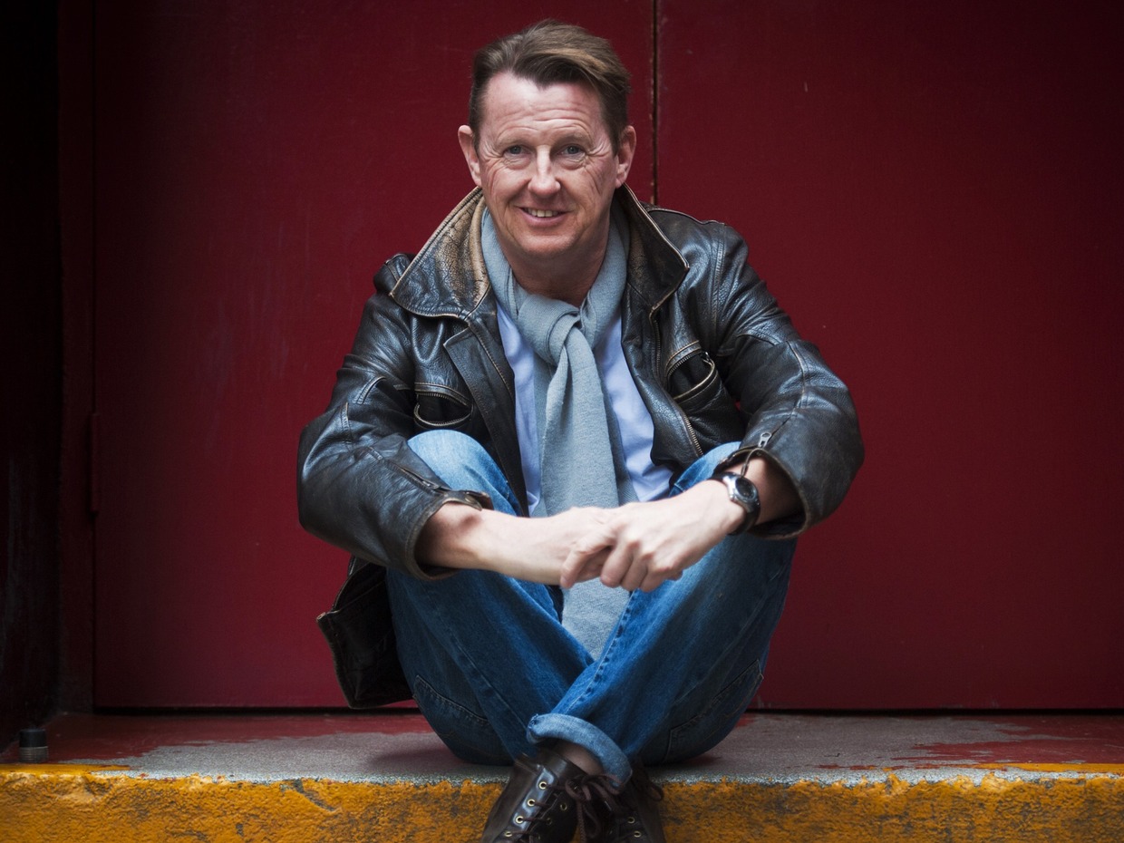 Kevin Kennedy in a brown leather jacket, grey scarf and jeans, sitting on a step in front of a red door and smiling