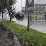 Killay Square under floodwater in heavy rain, taped off with red and white barrier tape, a car stranded in the water by the junction