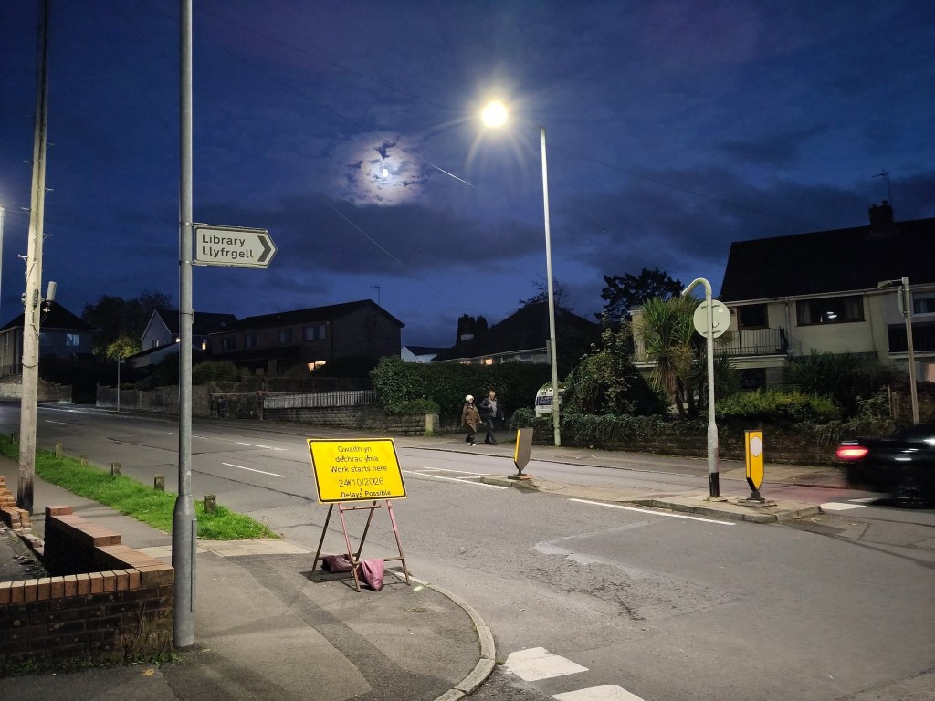 A yellow roadworks sign reading Work starts here 24/10/2026, delays possible, at dusk on Goetre Fawr Road in Killay, beside a sign pointing to the library