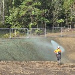 Two workers in orange hi-vis and hard hats spraying a green seed mix from a hose onto a bare earth bank beside temporary fencing, with woodland behind
