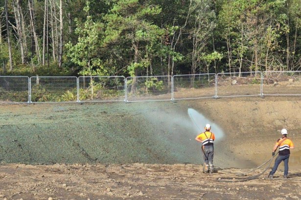 Two workers in orange hi-vis and hard hats spraying a green seed mix from a hose onto a bare earth bank beside temporary fencing, with woodland behind