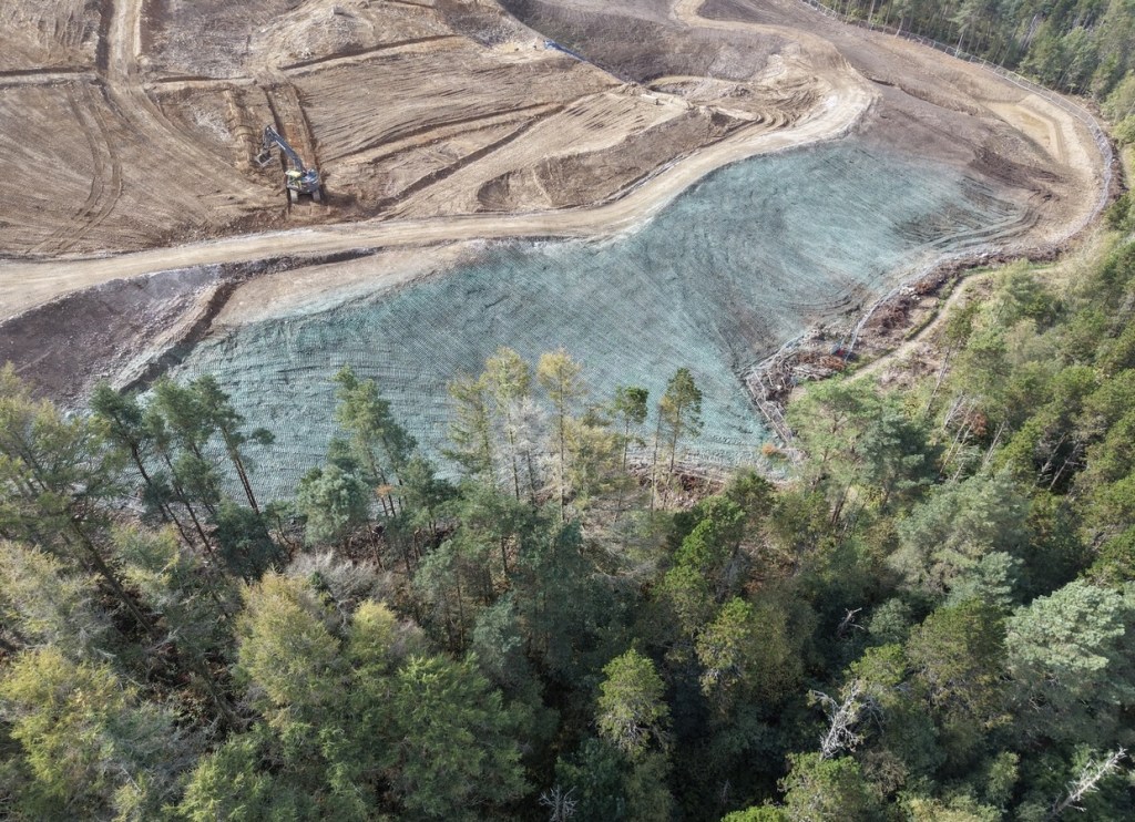 Drone view of a large blue-green hydroseeded slope on Kilvey Hill, with brown earthworks and a digger above it and conifer woodland below