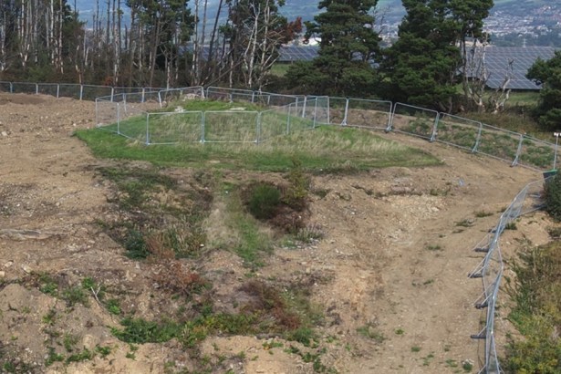 A grassy mound ringed by temporary metal fencing on bare, dug-over ground on Kilvey Hill, with conifers behind and solar panels and Swansea in the distance