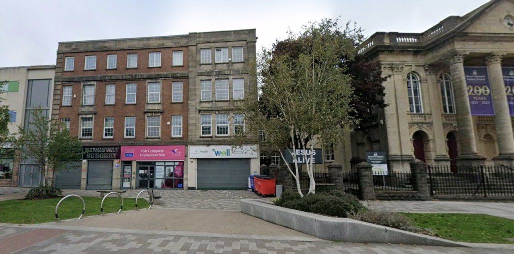 Gower College Swansea's Hyb Cyflogaeth Employment Hub on the ground floor of a red brick building on the Kingsway in Swansea, between Kingsway Surgery and a Well pharmacy