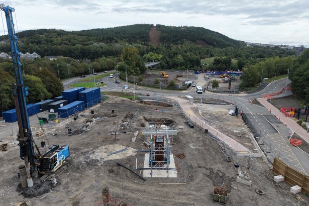 Drone view of the Skyline gondola base station site at Landore, with a piling rig, blue site cabins and a scaffolded column of steel reinforcement on a concrete base, and the cleared gondola line running up Kilvey Hill behind