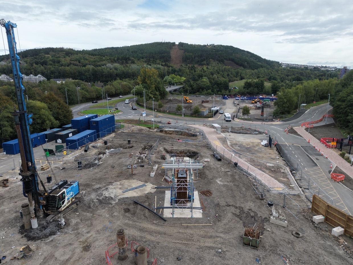 Drone view of the Skyline gondola base station site at Landore, with a piling rig, blue site cabins and a scaffolded column of steel reinforcement on a concrete base, and the cleared gondola line running up Kilvey Hill behind