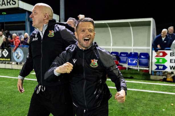 Lee John in a black Llanelli Town tracksuit top celebrating with a clenched fist on the pitch at night, with a colleague beside him and the dugout behind