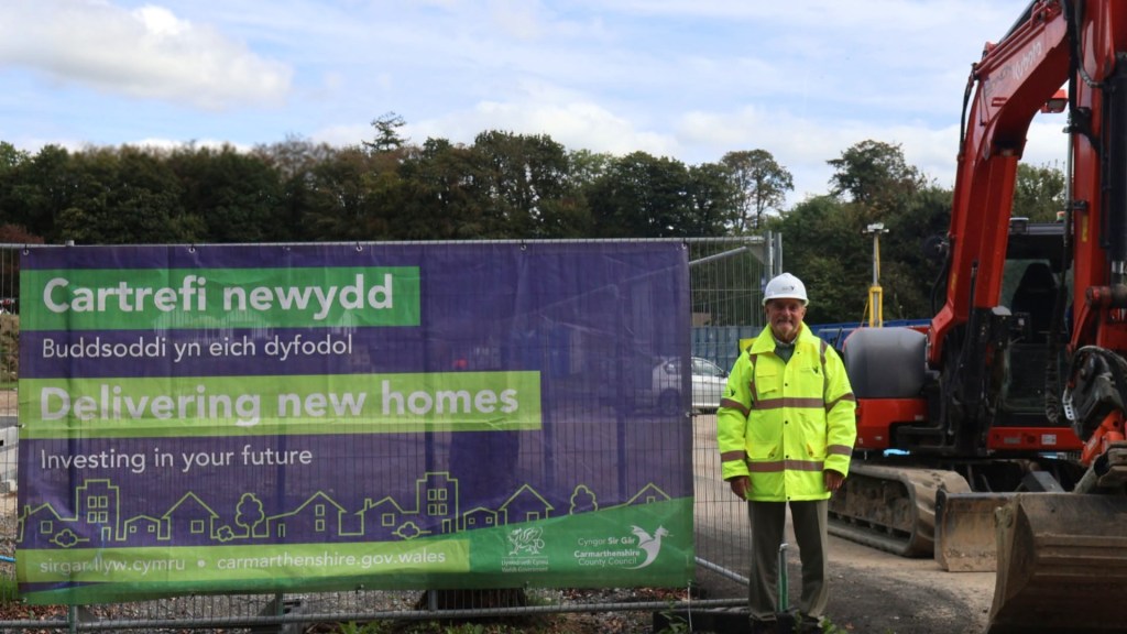 Cllr Emlyn Schiavone in a hi-vis jacket and hard hat beside a digger and a council banner reading Delivering new homes at the Cilycwm Road site in Llandovery