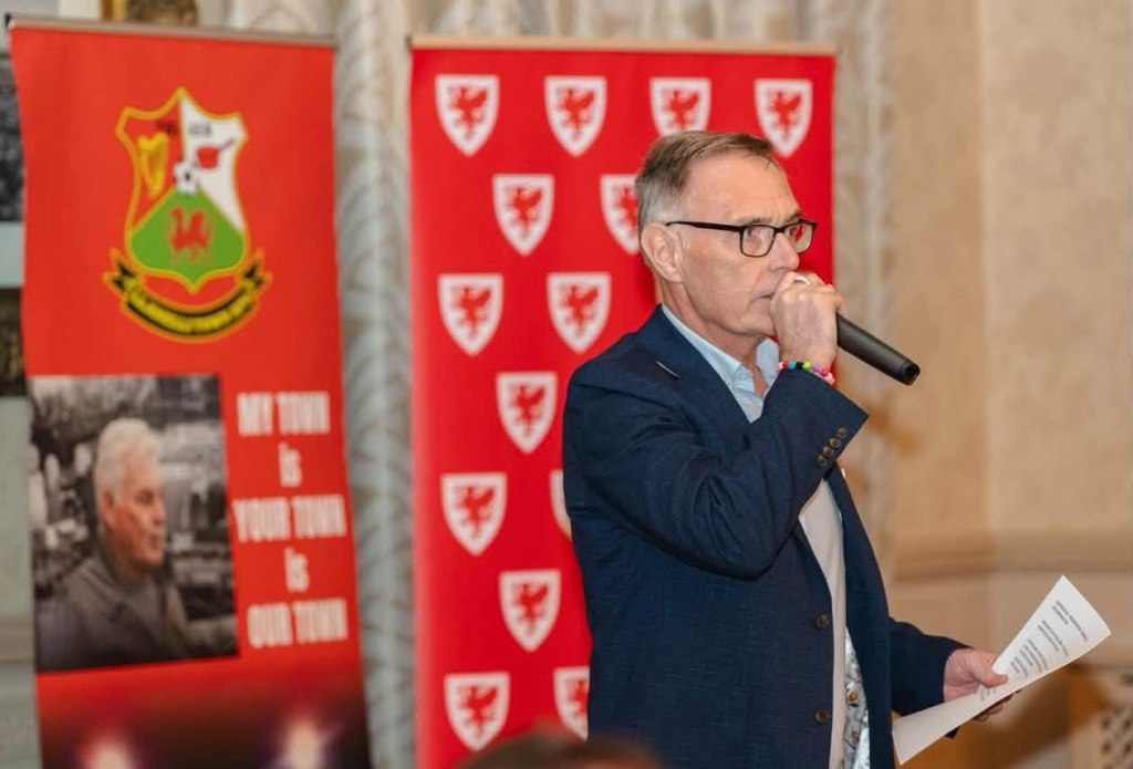 Colin Staples speaking into a microphone in front of Llanelli Town and FAW banners