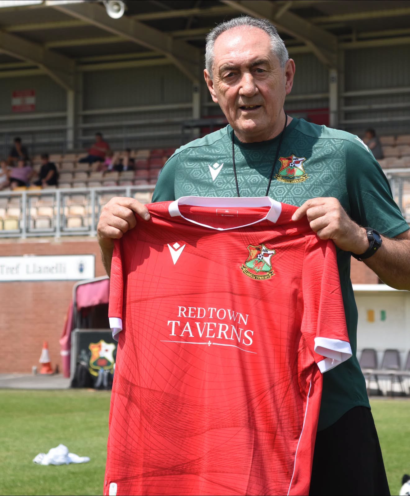 Terry Boyle in a green Llanelli Town training top holding up a red Llanelli Town home shirt at Stebonheath Park