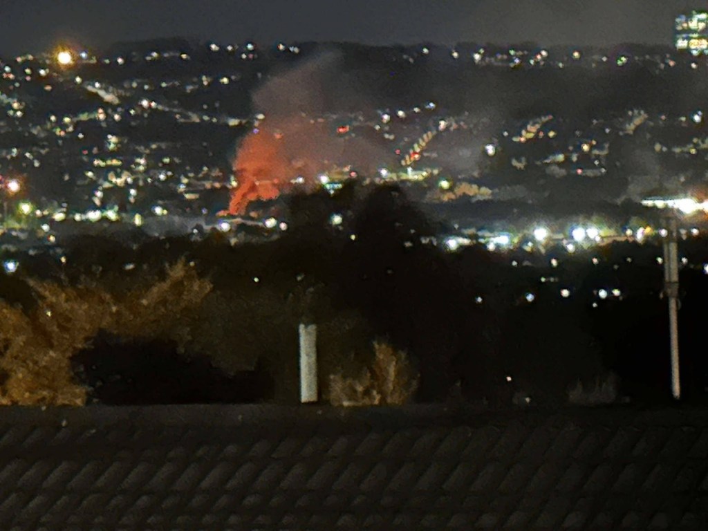 Orange flames and a column of smoke rising at night among the street lights of Llansamlet, seen from a rooftop