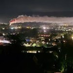 A long plume of white smoke, lit orange at its base, stretching across the night sky above the lights of Llansamlet