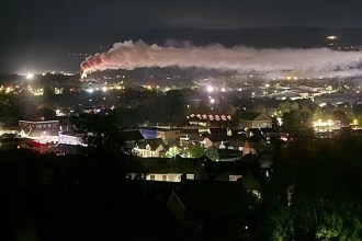 A long plume of white smoke, lit orange at its base, stretching across the night sky above the lights of Llansamlet