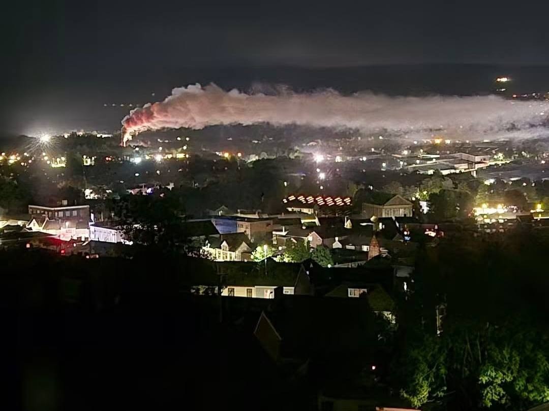 A long plume of white smoke, lit orange at its base, stretching across the night sky above the lights of Llansamlet