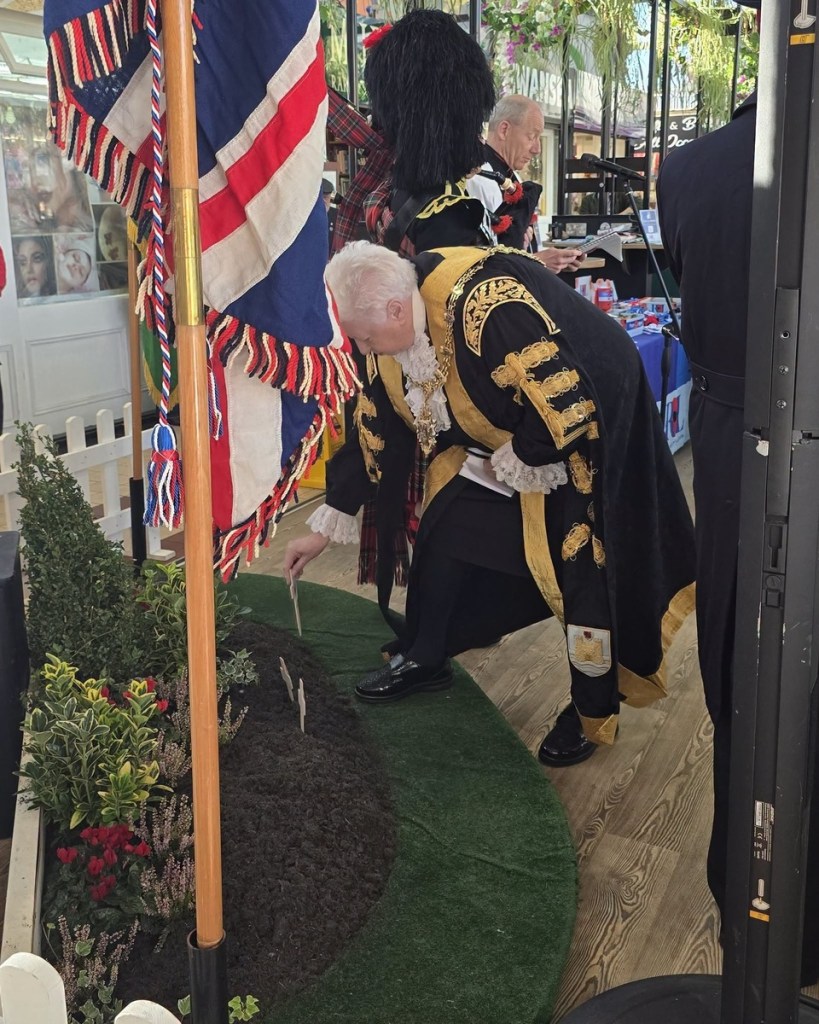 The Lord Mayor of Swansea in black and gold robes bends to place a small wooden cross in a garden of remembrance beside a Union flag.