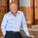 Lyndon Faulkner, smiling, in a light blue shirt, leaning on a dark counter in an office with wooden panelling