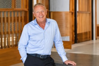 Lyndon Faulkner, smiling, in a light blue shirt, leaning on a dark counter in an office with wooden panelling