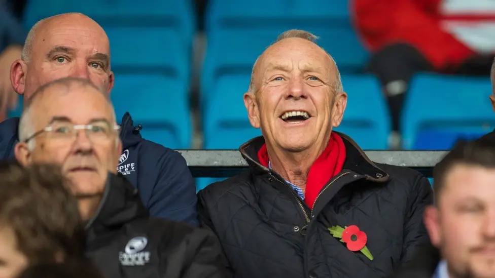 Lyndon Faulkner, in a red scarf and dark quilted jacket with a poppy, laughing in the stand at Cardiff Arms Park