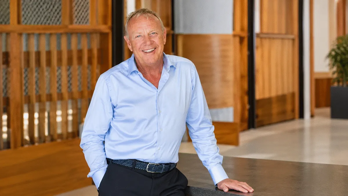 Lyndon Faulkner, smiling, in a light blue shirt, leaning on a dark counter in an office with wooden panelling