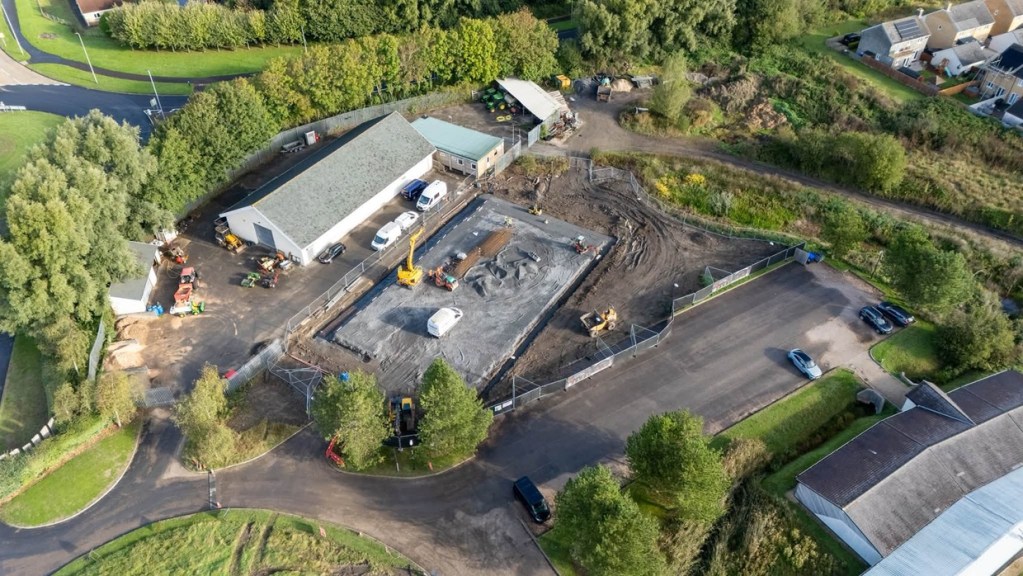 Aerial view of a fenced building site at Machynys with a grey concrete slab, diggers and vans beside a maintenance shed and car park